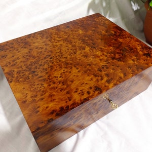 Puede incluir: Una caja de madera con un acabado de madera de raíz y una cerradura de latón. La caja está cerrada y tiene una superficie lisa y pulida.