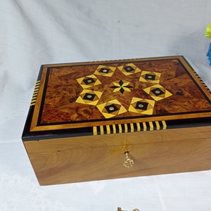 Puede incluir: Una caja de madera con una incrustación decorativa de formas geométricas en tonos marrones, negros y amarillos. La caja tiene un borde rayado negro y amarillo y una cerradura de latón.