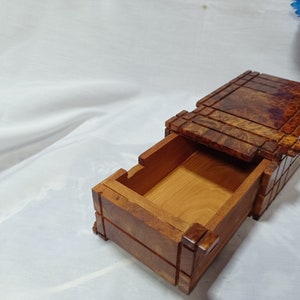 Peut inclure: Une boîte en bois avec un tiroir coulissant. La boîte est en bois foncé avec un motif de grain naturel. Le tiroir est ouvert et révèle un intérieur en bois clair.