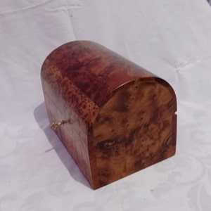 Pode incluir: Uma pequena caixa de madeira com uma tampa curva e uma fechadura. A caixa é feita de madeira escura com um tom marrom avermelhado e um padrão de grão único.