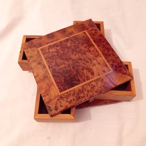 Pode incluir: Uma caixa de madeira com um design em camadas. A camada superior é uma caixa quadrada com uma borda decorativa. A caixa é feita de madeira marrom clara com uma incrustação de madeira marrom escura.