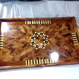 Pode incluir: Uma bandeja retangular de madeira com um design de incrustação geométrica. A bandeja tem duas alças e um acabamento brilhante. A incrustação é feita de madeiras de diferentes cores, incluindo preto, branco e castanho.