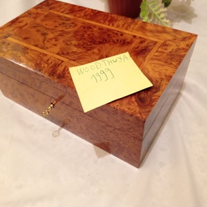 Puede incluir: Una caja de madera con una cerradura y una nota adhesiva amarilla que dice "WOODTHUYA 1999".