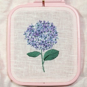May include: A purple and white embroidered hydrangea flower with green leaves in a pink embroidery hoop.