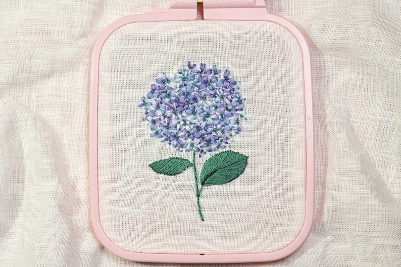 May include: A purple and white embroidered hydrangea flower with green leaves in a pink embroidery hoop.
