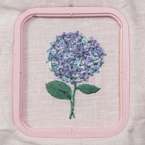 May include: A purple and white embroidered hydrangea flower with green leaves in a pink embroidery hoop. The hoop is labeled "100 x 100 mm".