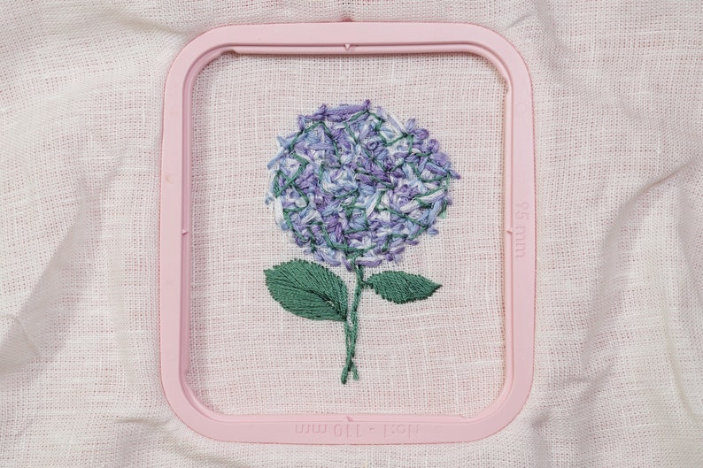 May include: A purple and white embroidered hydrangea flower with green leaves in a pink embroidery hoop. The hoop is labeled "100 x 100 mm".