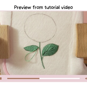 May include: A close-up of a white fabric with a green embroidered flower. The flower has a circle in the center and two leaves. The fabric is stretched over a wooden embroidery hoop.