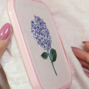 May include: A close-up of a pink embroidery hoop with a white fabric inside. The fabric has a purple and green embroidered flower design.