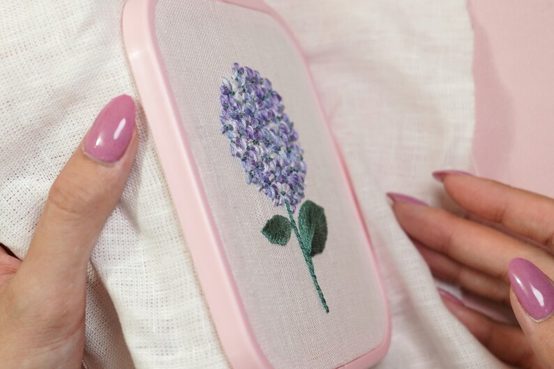 May include: A close-up of a pink embroidery hoop with a white fabric inside. The fabric has a purple and green embroidered flower design.