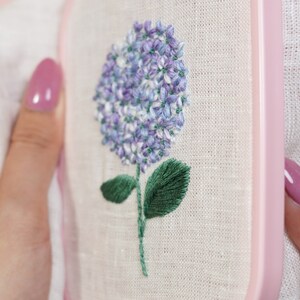 May include: A close-up of a hand-stitched embroidery of a purple hydrangea flower with green leaves on a white linen fabric. The embroidery is framed in a pink wooden hoop.