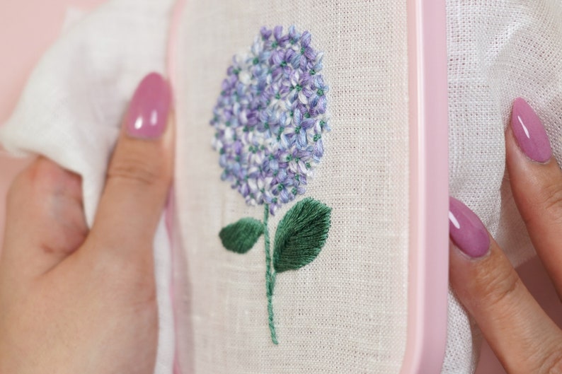 May include: A close-up of a hand-stitched embroidery of a purple hydrangea flower with green leaves on a white linen fabric. The embroidery is framed in a pink wooden hoop.