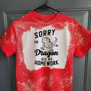 Peut inclure: T-shirt rouge avec un centre blanc blanchi. Le t-shirt présente le texte "SORRY THE Dragon ATE MY HOMEWORK" en noir, ainsi qu'une illustration de dessin animé d'un dragon.