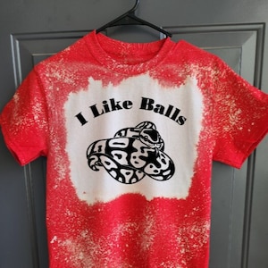 May include: Red t-shirt with a white bleach design. The design features a snake with the text "I Like Balls" above it.