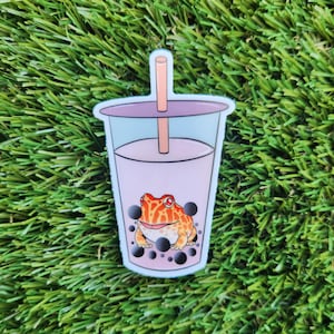 May include: A sticker of a pink and purple bubble tea with a frog inside and black boba pearls. The frog is orange and brown with red eyes.