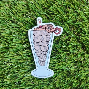 May include: A sticker of a cartoon snake in a milkshake glass with a straw. The snake is brown and white with a red tongue and a heart-shaped smile. The milkshake glass is blue and white.