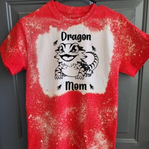 Peut inclure: T-shirt rouge avec un motif éclaboussé d'eau de Javel. Le t-shirt présente une image de dessin animé en noir et blanc d'un dragon barbu avec le texte "Dragon Mom" en dessous.