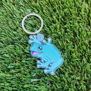 May include: A blue frog keyring with a gold metal ring. The frog is cartoon-style and has a light blue body with darker blue spots. The frog is sitting on its hind legs with its front legs raised.