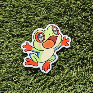 May include: A sticker depicting a cartoon frog with large eyes and an open mouth. The frog is green with orange, red, and blue details. The sticker is placed on a green, textured surface.