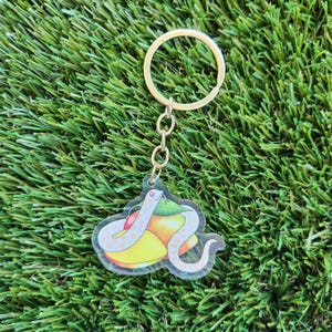 May include: A white and orange snake keychain with a gold chain. The snake is curled around a yellow and orange fruit.