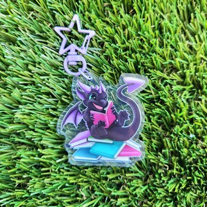 May include: A black dragon with purple wings is reading a pink book while sitting on a stack of colorful books. The dragon is a charm on a clear keychain with a purple star-shaped clasp.