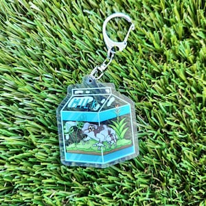 May include: A clear plastic keychain shaped like a milk carton with a cartoon frog inside. The frog is brown and white with a blue background. The keychain says "MILK" on the front.
