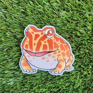May include: A cartoon sticker of a red and orange frog with white spots. The frog has a wide smile and red eyes.