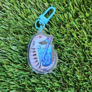May include: A clear keychain featuring a cartoon gecko enjoying a blue boba tea with a blue and orange straw. The gecko is light brown with dark brown stripes. The boba tea contains white boba pearls.
