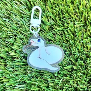 May include: White snake keychain with blue eyes and a red tongue. The keychain is made of clear acrylic and has a white metal clasp.