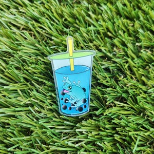 May include: A blue and green frog is inside a clear plastic cup with a blue liquid and black boba pearls. A green straw is sticking out of the top of the cup.