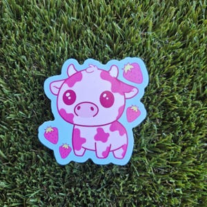 May include: A sticker featuring a cartoon cow with pink spots and a pink body. The cow has large, round eyes and is surrounded by strawberries. The sticker has a light blue border and is set against a green background.