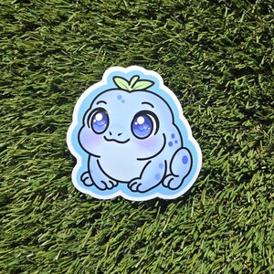 May include: A die-cut sticker depicting a cartoon frog-like character in blue hues. The creature has large eyes, a small smile, and a leaf detail on its head. The sticker is outlined in white, set against a green background, and is suitable for various applications.