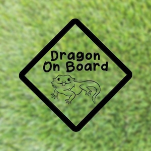 May include: A black diamond-shaped sign featuring the words "Dragon On Board" and a cartoon lizard. The sign is set against a blurred green background, resembling grass.