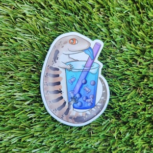 Peut inclure: Un autocollant représentant un lézard de dessin animé buvant du bubble tea bleu avec une paille violette. Le lézard est brun et blanc avec un motif rayé. Le bubble tea contient des perles de tapioca blanches.