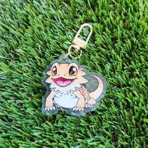 May include: A clear keychain with a cartoon image of a bearded dragon. The dragon is orange and brown with a white belly and is smiling. The keychain has a gold metal ring.