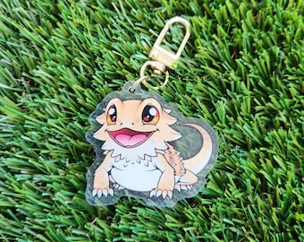 May include: A clear keychain with a cartoon image of a bearded dragon. The dragon is orange and brown with a white belly and is smiling. The keychain has a gold metal ring.