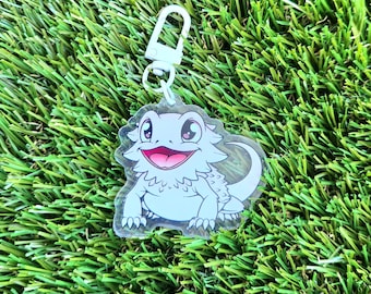 May include: A white bearded dragon keychain with a light blue background. The dragon has a pink tongue and pink eyes. The keychain is made of clear acrylic.