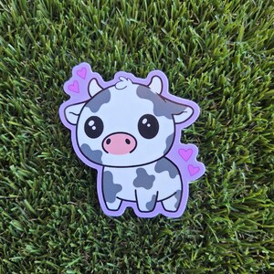 May include: A cute cartoon cow sticker with a white body, gray spots, and a pink nose. The cow has large black eyes and is outlined in purple. Three small purple hearts are on the side.