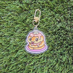 Peut inclure: Un porte-clés en acrylique transparent avec un motif de crêpes et un personnage de serpent mignon, garni de beurre et de sirop. Le porte-clés a un fermoir doré et un petit anneau. Le design est souligné en rose. Le fond est vert.