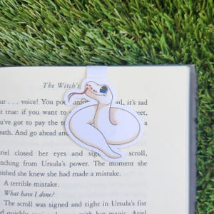 May include: A white snake bookmark with blue eyes and a red tongue. The bookmark is curled up and has a magnetic clip on the back.