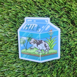 May include: A sticker of a milk carton with a frog inside. The carton is blue and white with the word "MILK" printed on the front. The frog is green and white with black spots. There are green plants inside the carton.