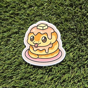 Peut inclure: Autocollant représentant une pile de crêpes avec un visage de serpent. Les crêpes sont jaunes avec du sirop et un carré de beurre. Le serpent a des yeux bruns et une langue rose. L'autocollant est sur une assiette rose.