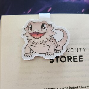 May include: A white bookmark with a cartoon image of a light brown bearded dragon with big eyes and a wide open mouth. The dragon is sitting on its hind legs with its front paws in the air. The bookmark is attached to a white metal clip.