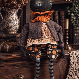 May include: A fabric doll with a round black pumpkin head, blue eyes, and an orange ruffled collar. It wears a brown herringbone jacket over a cream-coloured top with a leaf print, pumpkin-patterned trousers, and black and grey striped legs.