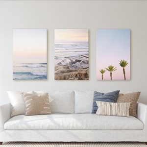 Puede incluir: Tres impresiones enmarcadas de escenas de playa. La impresión de la izquierda muestra un océano azul con olas blancas y un cielo rosa y morado. La impresión del medio muestra una playa rocosa con un océano azul y un cielo rosa y morado. La impresión de la derecha muestra tres palmeras contra un cielo rosa y morado.