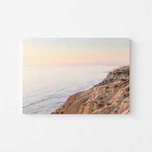 Puede incluir: Una vista panorámica de un acantilado arenoso que domina un vasto océano con suaves olas. El cielo es de un suave rosa pálido y azul, creando una atmósfera serena y tranquila.