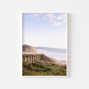 Puede incluir: Un puente de hormigón sobre una playa de arena con olas azules del océano rompiendo en la costa. El puente está bordeado de hierba verde y flores silvestres. El cielo es de un azul claro con nubes blancas.