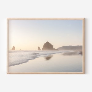 May include: A scenic photograph of a rocky coastline with a large rock formation in the distance. The water is calm and reflects the sky, which is a soft blue with hints of pink and orange. The beach is sandy and appears to be empty.
