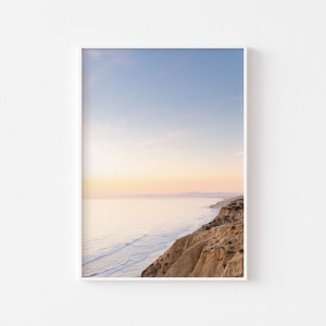 Puede incluir: Una vista panorámica de un acantilado arenoso que domina un océano azul con olas blancas rompiendo en la costa. El cielo es de un azul suave con una puesta de sol rosa y naranja en la distancia.