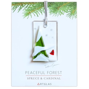 Peut inclure: Une décoration en verre fusionné rectangulaire représentant un sapin vert avec des accents de neige blanche et un petit cardinal rouge sur fond blanc. L'ornement est suspendu par un ruban argenté sur une carte marquée PEACEFUL FOREST.